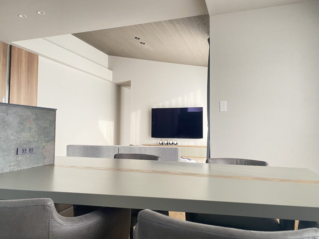 グレーのダイニングテーブルに４脚の椅子が並ぶ様子。奥にはソファとテレビがある様子が撮影されたグレー基調の落ち着いた雰囲気のリビング写真。