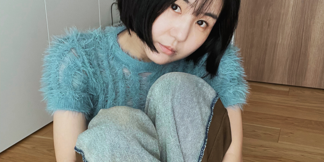 淡いブルーのふわっとしたトップスを着て、床に座り膝を抱える女性の姿。少し見上げるような視線が印象的で、静かな内面を感じさせる一枚。