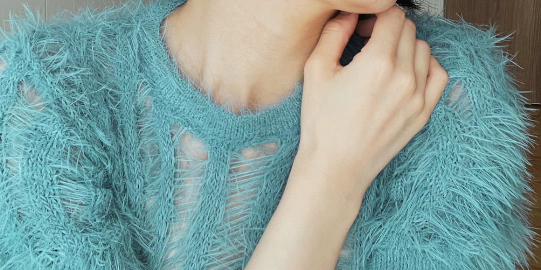 淡いブルーの透け感のあるトップス越しに、首元に手を添える仕草のクローズアップ。柔らかい質感と繊細な空気感が伝わるビジュアル。