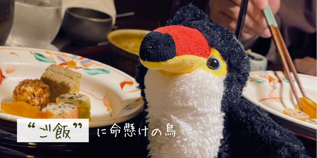 食事中のテーブルで、料理を前にしたオニオオハシのぬいぐるみ「ねぐち」が身を乗り出している様子。「“ご飯”に命懸けの鳥」という文字が添えられ、食べることへの熱意をユーモラスに表現している。