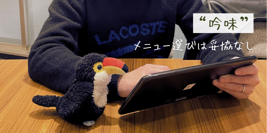 タブレットでメニューを選ぶ人物の隣で、オニオオハシのぬいぐるみ「ねぐち」が真剣な表情で見守っている。「“吟味” メニュー選びは妥協なし」という文字が添えられ、食事選びへのこだわりをコミカルに表現している。